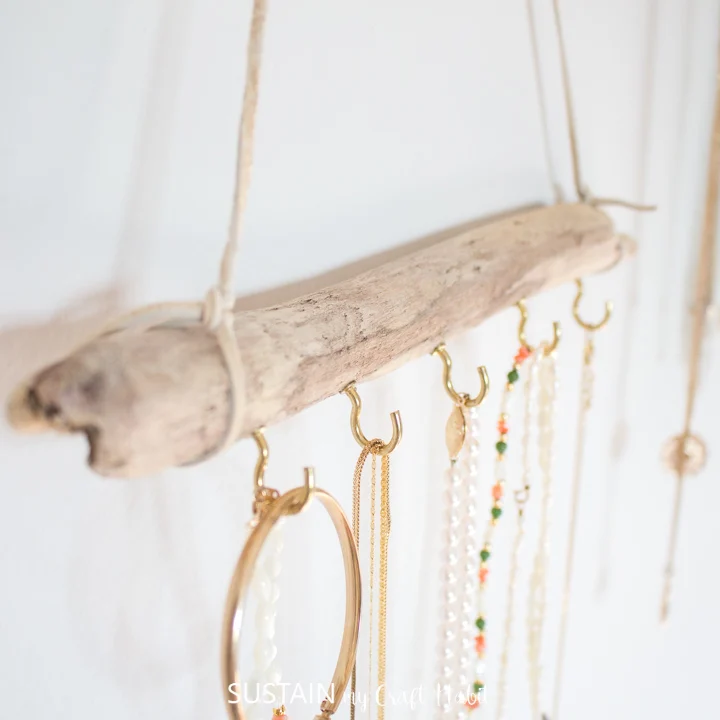 Homemade jewellery display idea with a long piece of driftwood and gold cup hooks. Various necklaces and bracelets hang on the hooks.