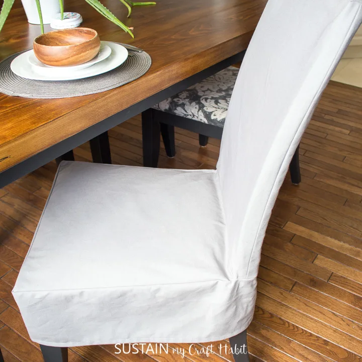 A completed dining chair slipcover on a wooden chair in a dining room with wood flooring. A light gray canvas fabric was used to make the chair cover.