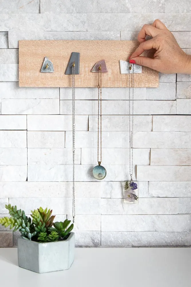 A wood jewelry display hanging on a white brick wall with necklaces placed on the hooks.