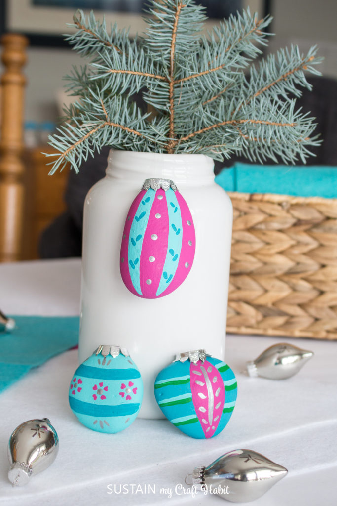 Christmas ornament painted rocks set against Christmas ornaments and a vase with pines.