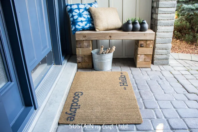 Porch with wooden bench, pillows, decor and a painted welcome mat.