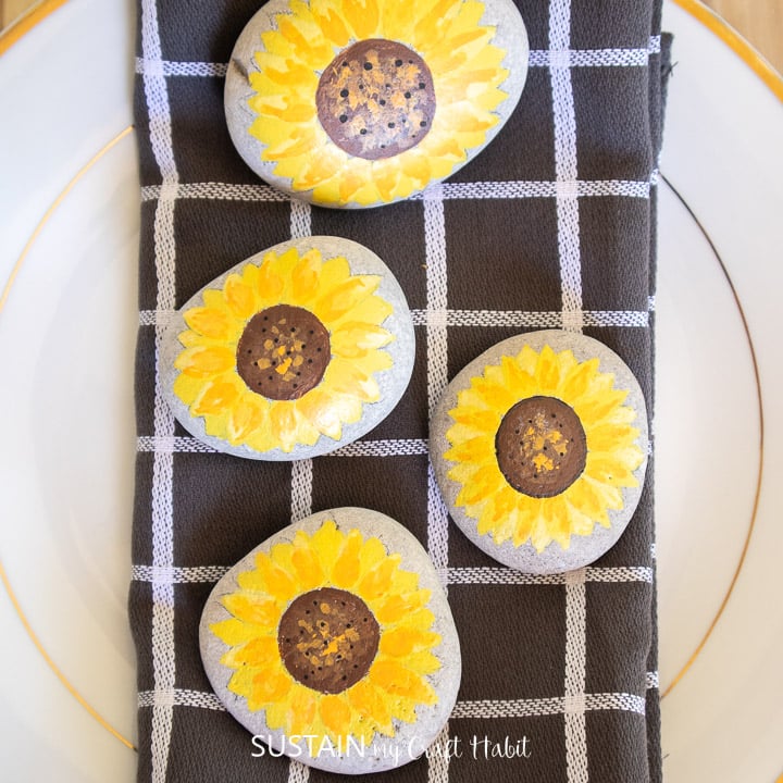 Four sunflower painted rocks on a dark gray checkered tea towel. 