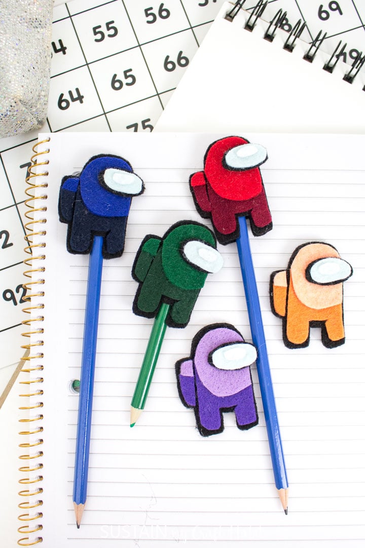 Felt Among us pencil toppers in blue, red, green purple and orange displayed on a lined school book.