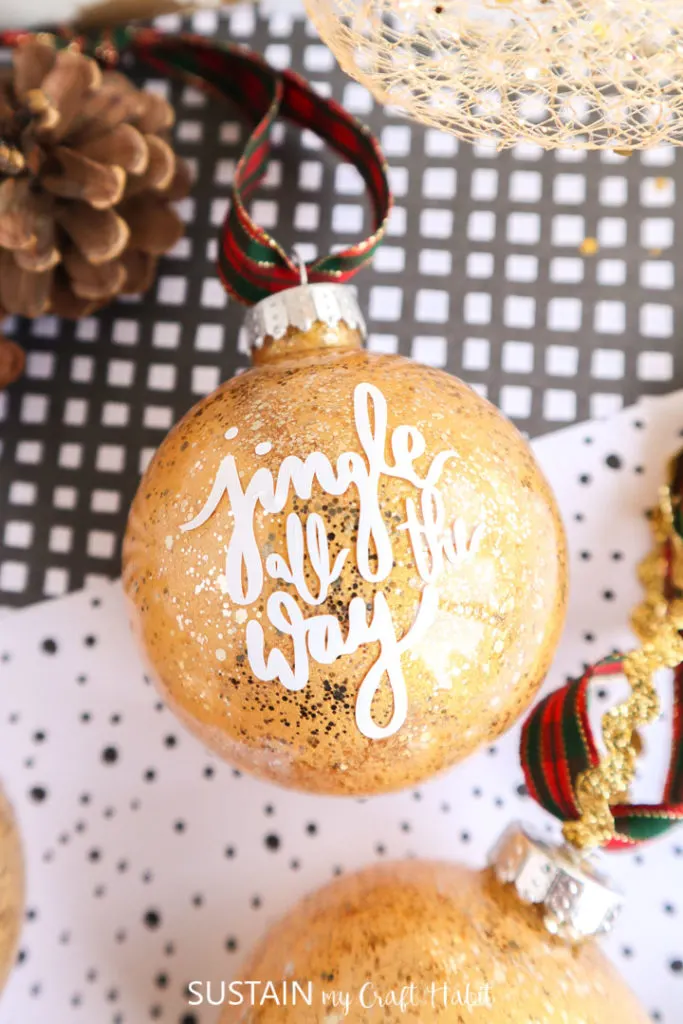 Close up of the vinyl words "jingle all the way" on the glittering gold painted glass ornament.