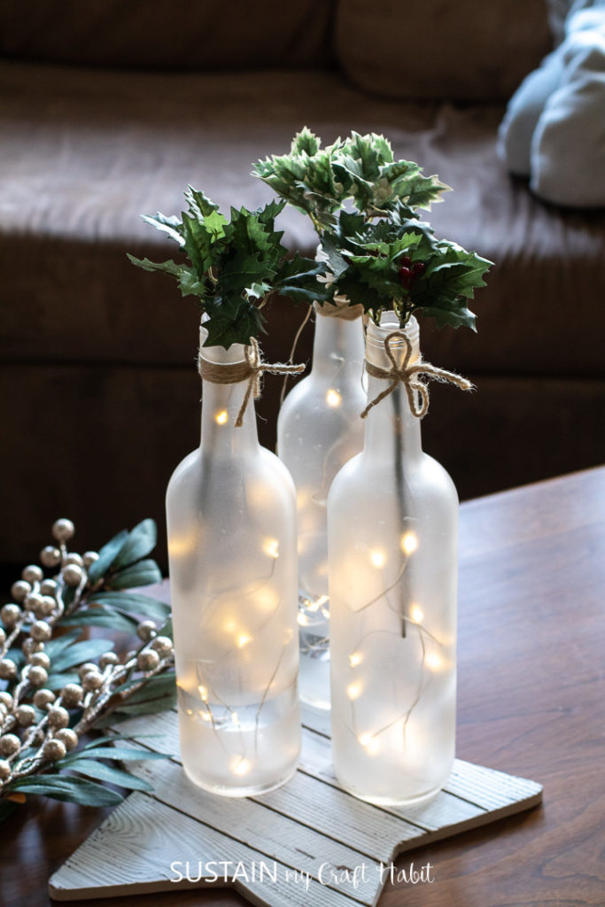 Frosted wine bottle centerpieces arranged with twinkle lights and fresh greenery.