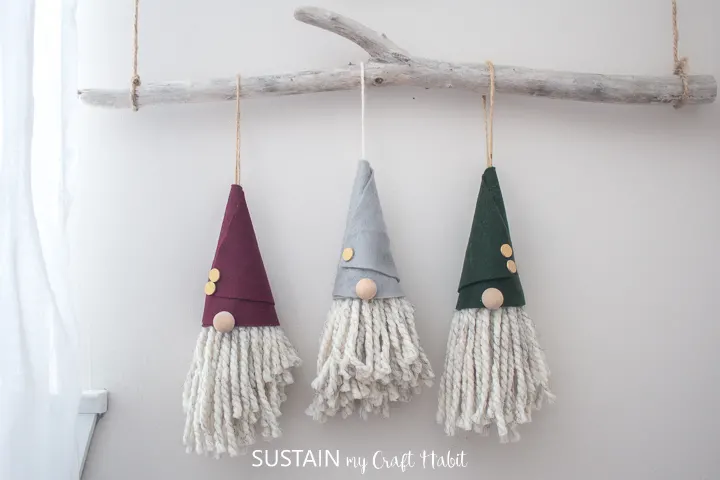 Three completed gnome christmas ornaments, hanging side by side on a branch of white-washed driftwood against a beige wall. 