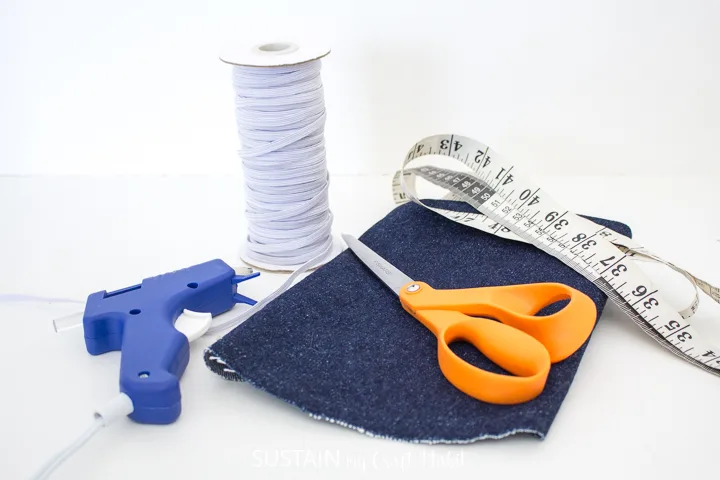 Materials needed to make elastic book bands including denim, measuring tape, scissors, elastic and a hot glue gun.