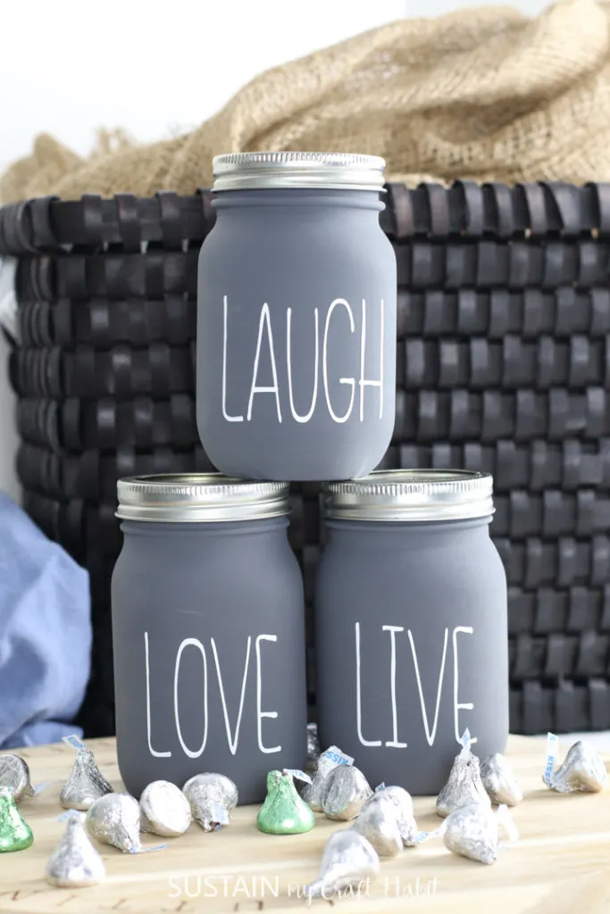 Finished chalky painted jars with lettering "laugh," "love," and "live" on the front. Placed next to a basket and chocolates.