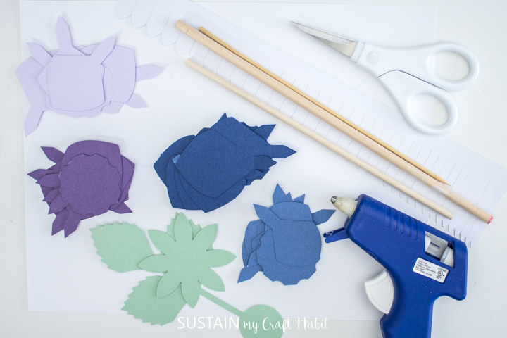 Matériel nécessaire pour assembler des fleurs de dahlia en papier, y compris du papier découpé, des brochettes, un pistolet à colle chaude et des ciseaux.