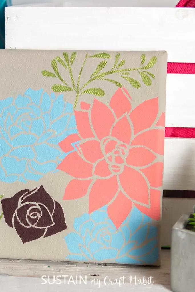 Canvas stencil art with flowers set on a shelf with other decor.