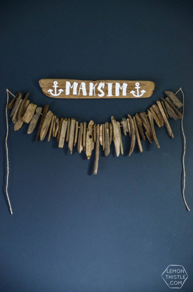 A painted piece of driftwood with a child's name hanging above a driftwood garland on a dark blue wall.