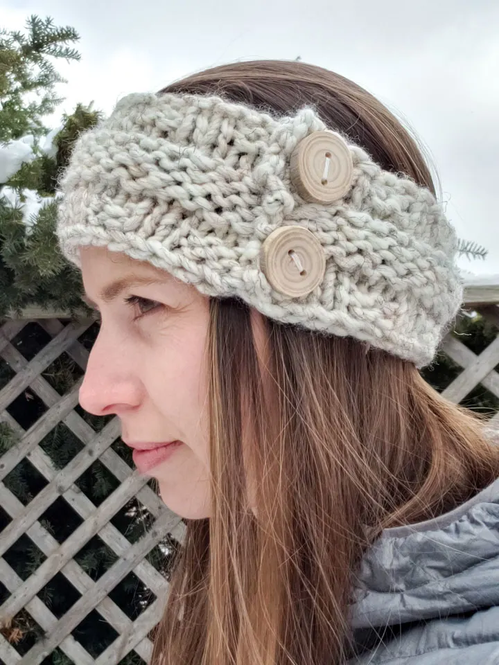 Side profile of a woman wearing a knitted headband made with cream wool yarn and two wood buttons for decoration.