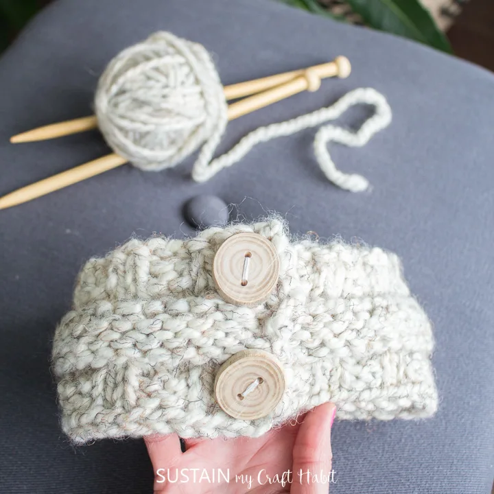 Holding a knitted headband made with cream chunky weight yarn two wooden buttons. A ball of yarn and wood straight knitting needles are in the background.