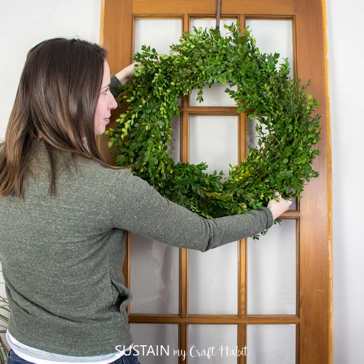 A woman hanging a boxwood wreath on a wooden door.
