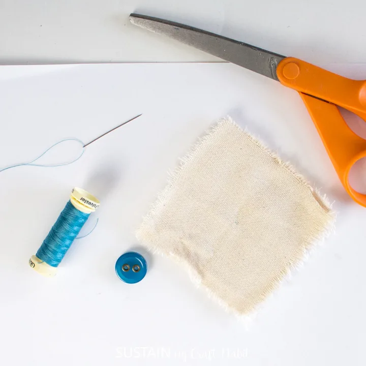 A needle with thread, blue button, canvas fabric square and scissors.