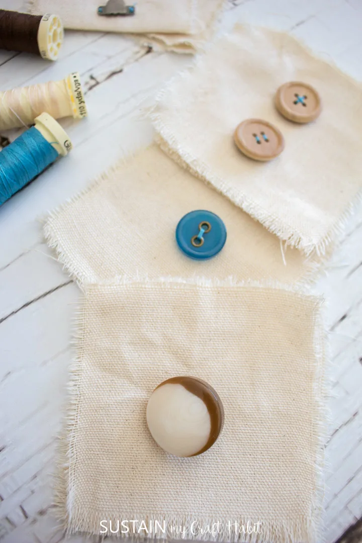 Different shaped buttons sewn to canvas fabric squares next to thread.