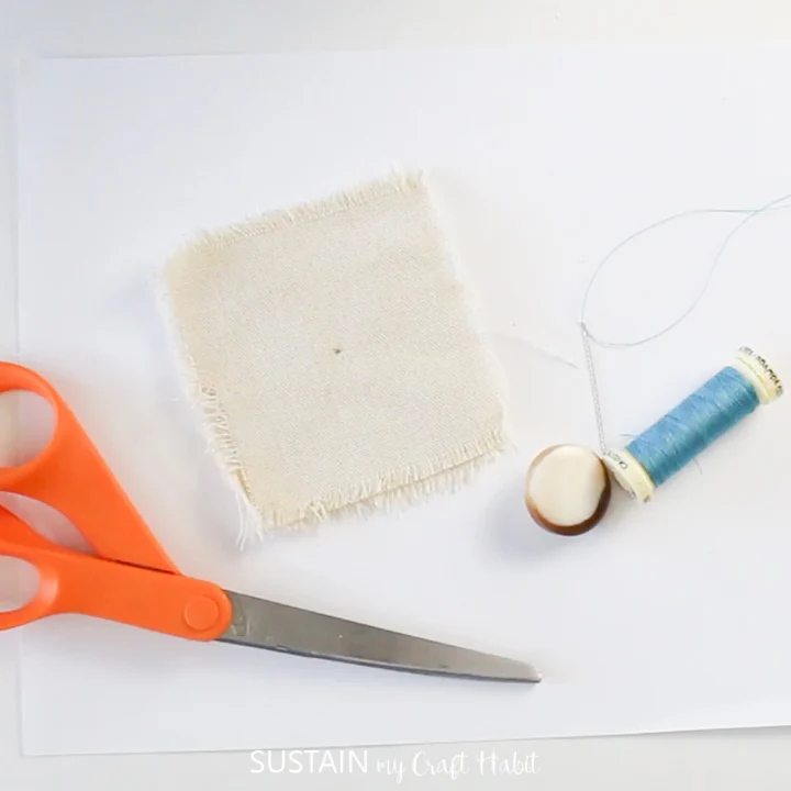 A needle with thread, blue button, canvas fabric square and scissors.