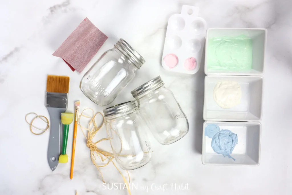 Materials needed to make decorative painted Easter jars including mason jars, paint, paintbrushes, rubber band and sandpaper.