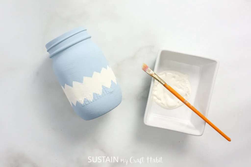 Paint brush and paint next to a mason jar painted with a zig zag design.