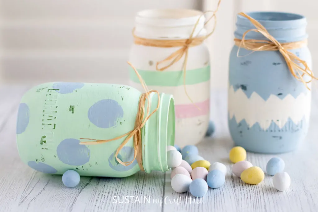 Three painted Easter jars wrapped in ribbon, one jar is laying on it's side with chocolate eggs spilling out.