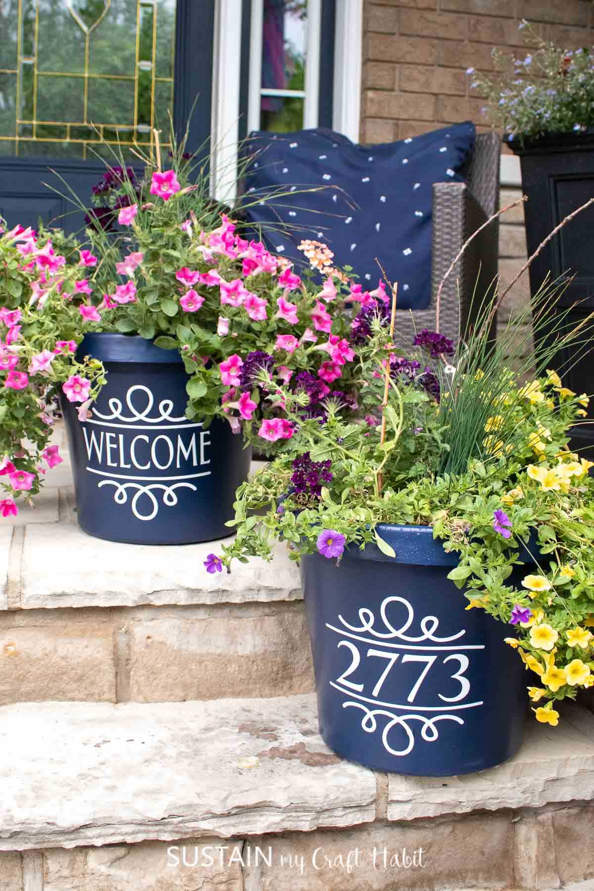 A pair of navy blue planters staggered on the steps of the porch. The planters are filled with colorful flowers and have white decals on the front. One decal says welcome and the other has a house number.