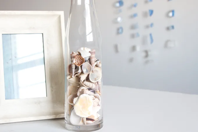 A picture frame next to a glass bottled filled with sea shells with a resin seaglass wall hanging in the background.