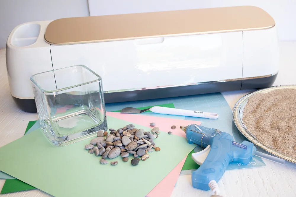 Materials needed to make a 3D paper cactus terrarium including a Cricut machine, cardstock paper, glass vase, sand, pebbles and hot glue.