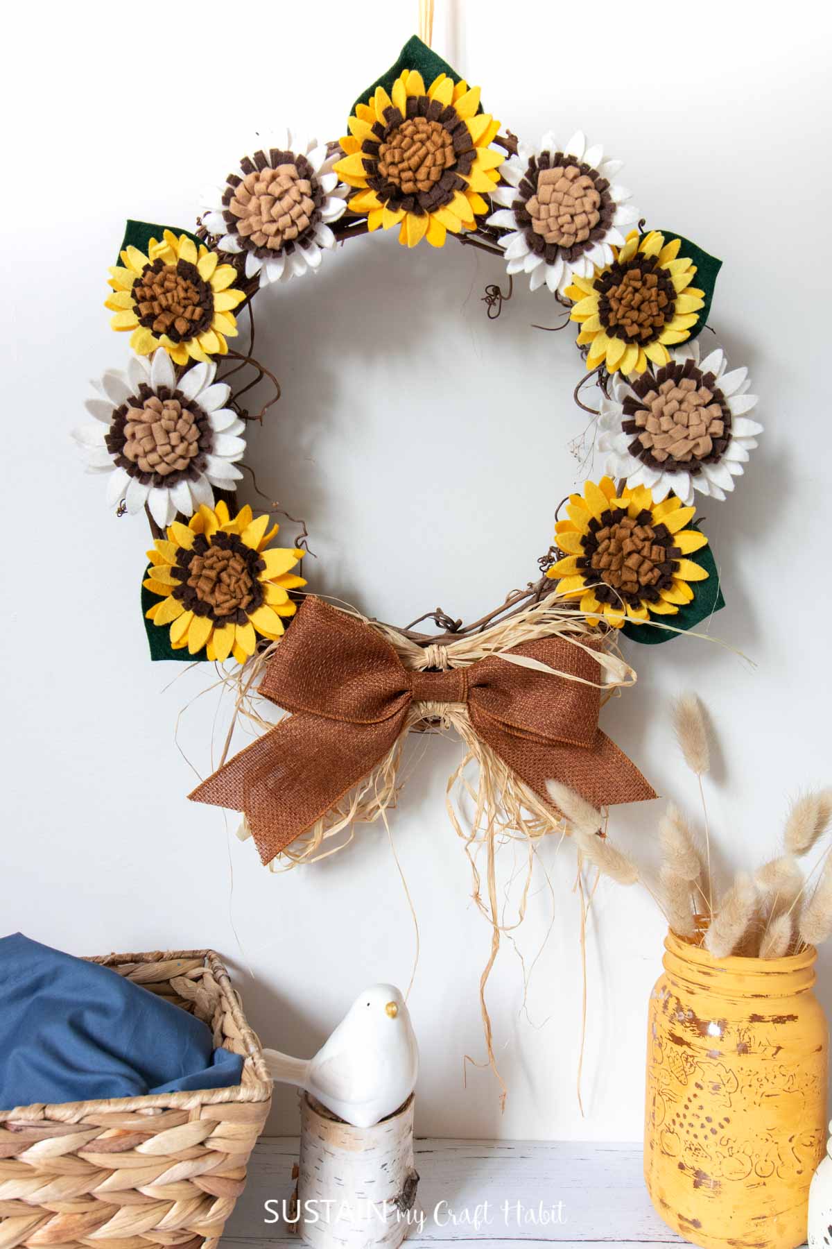 Felt DIY sunflower wreath hanging on a white wall. A yellow jar and white ceramic bird decorate the table underneath.