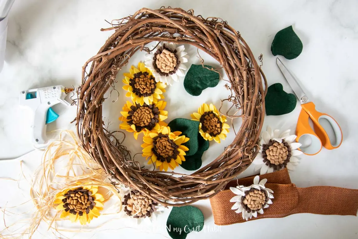 Materials needed to make a felt sunflower wreath including a grapevine wreath, felt flowers, scissors, ribbon and hot glue.