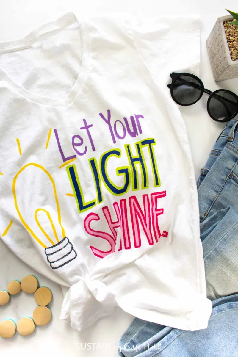 Tshirt painting with "let your light shine" in bright letters with a light bulb next to jeans and accessories.