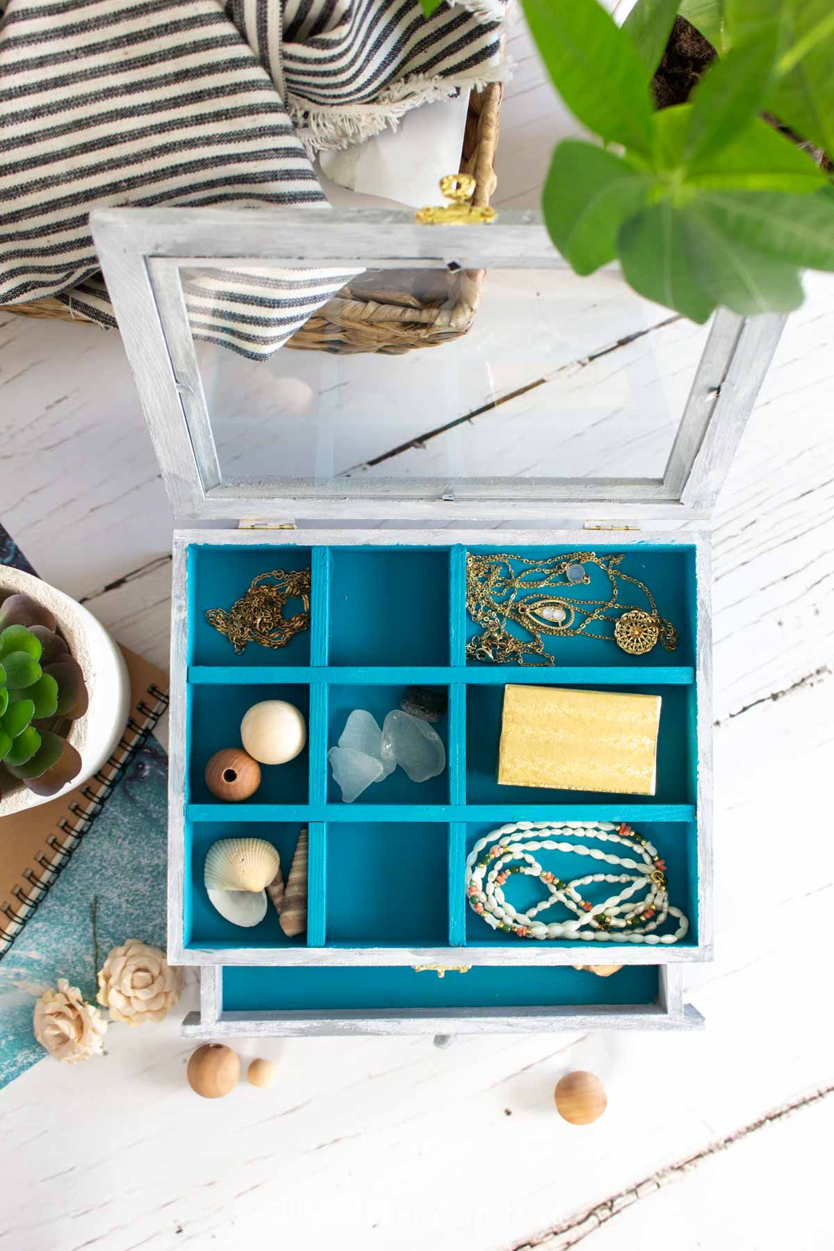 Overhead view of a hand painted jewelry box that's white washed on the outside and teal blue on the inside, filled with jewelry and trinkets.