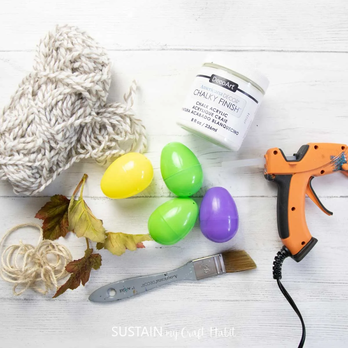 Materials needed for an upcycled plastic egg acorn ornament craft including plastic eggs, paint, hot glue, yarn, paintbrush and faux leaves.