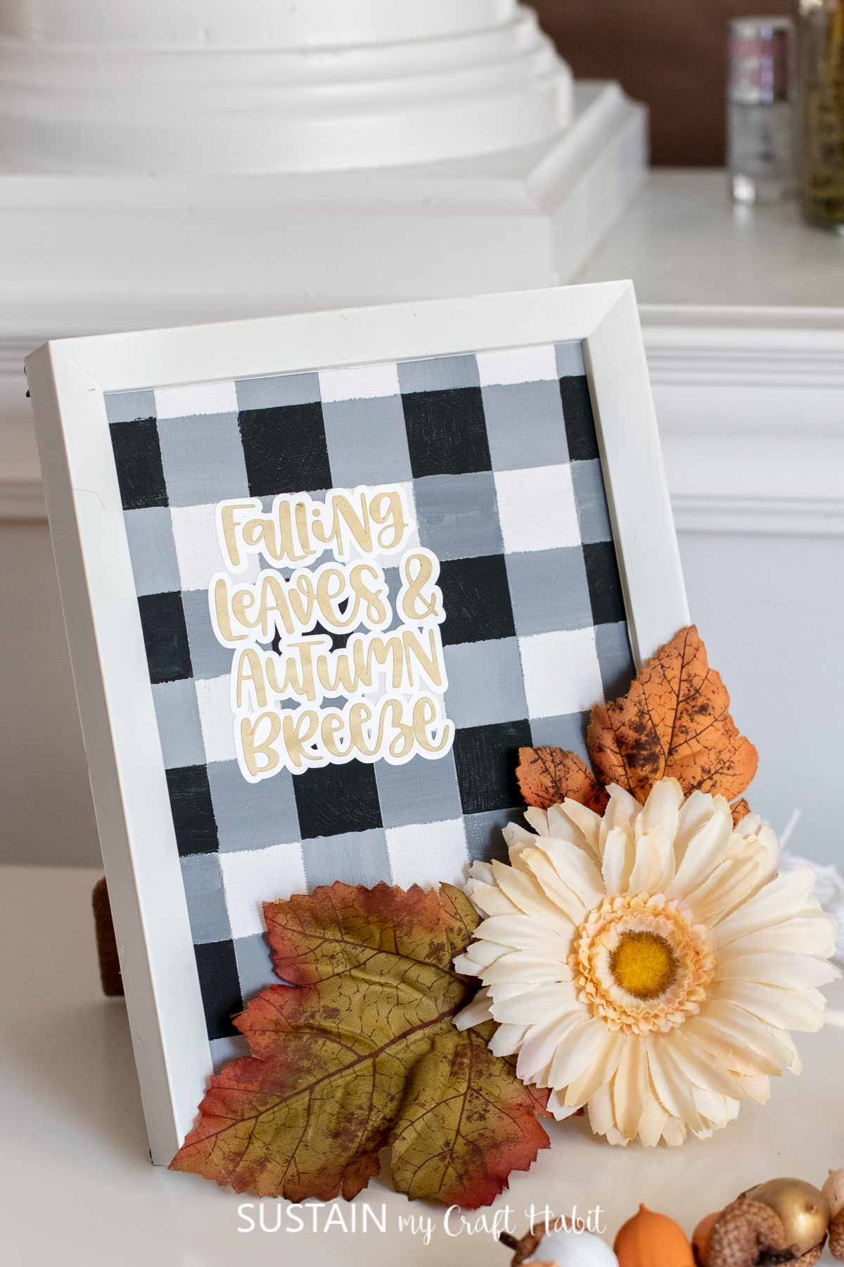 Painted buffalo plaid pattern in a frame with a "falling leaves and autumn breeze" cut out phrase and faux flowers and leaves.