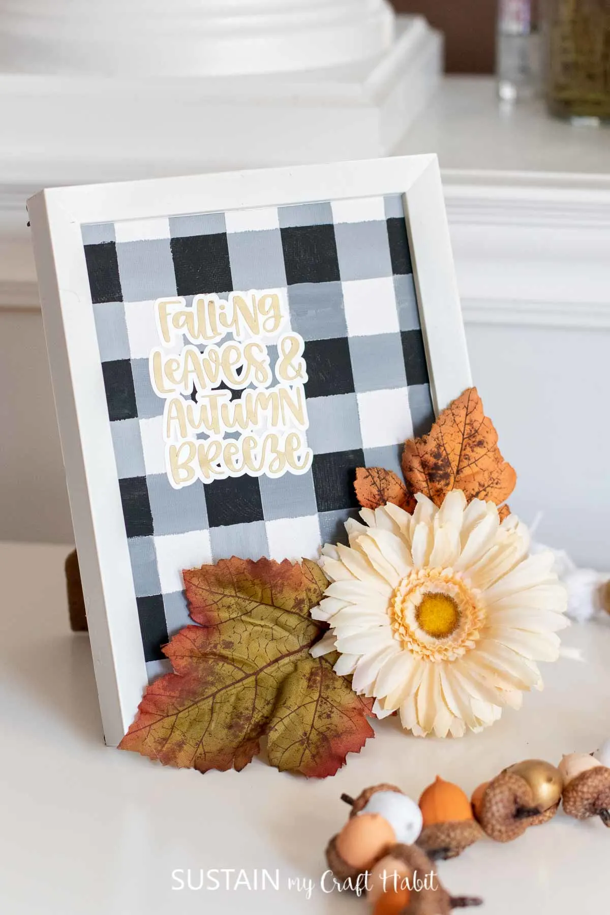 Painted buffalo plaid pattern in a frame with a "falling leaves and autumn breeze" cut out phrase and faux flowers and leaves.