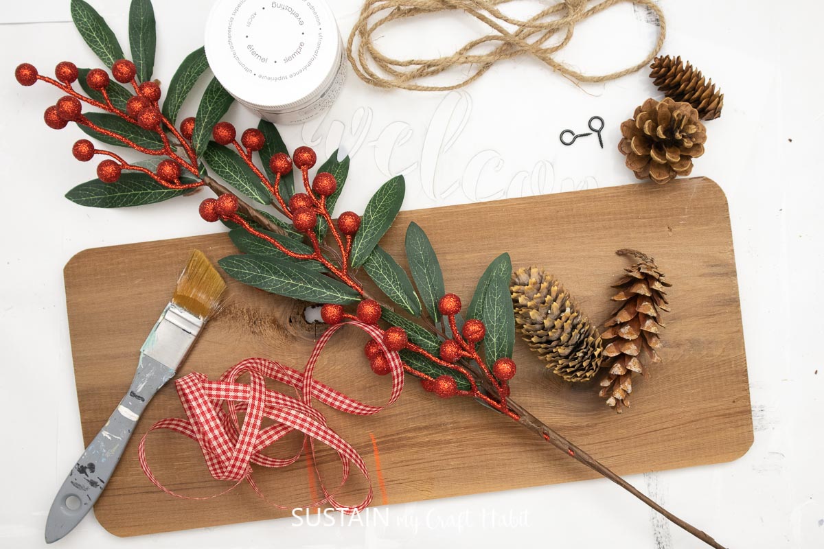 Materials needed to make a winter wood sign including a cedar plank, paint, paint brushes, eyelets, ribbon, twine and pine cones.