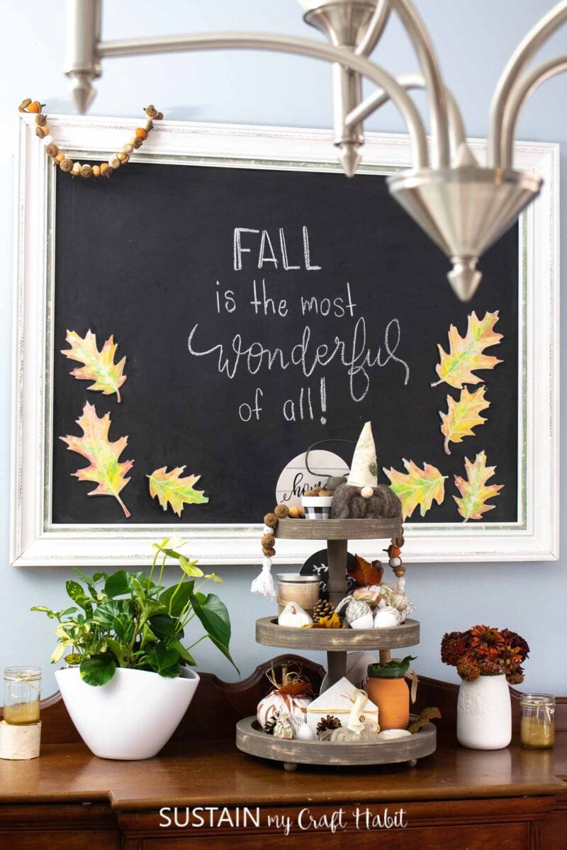 Framed chalkboard wall art decorated with leaves, acorns, and chalk writing placed next to a tired fall tray and vases.
