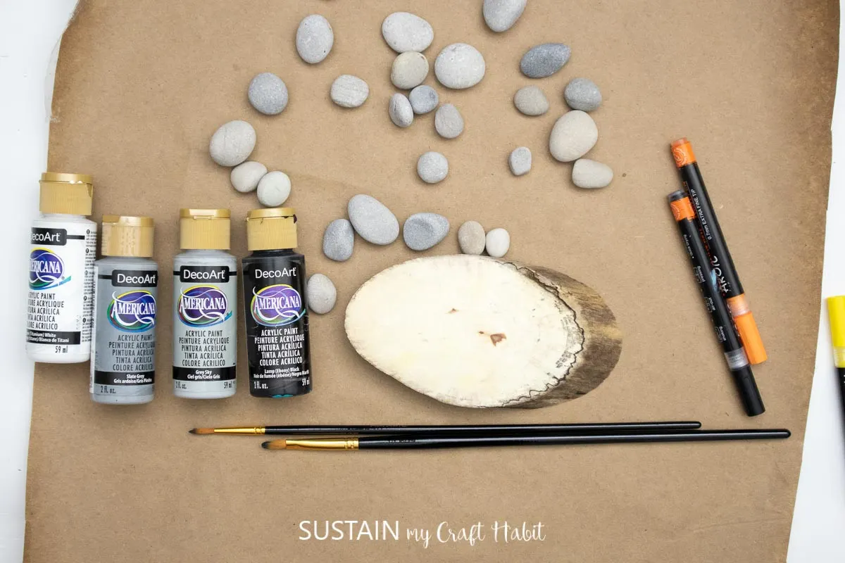 Materials needed to make a penguin pebble art craft including a wood slice, pebbles, paint, paint brushes and markers.
