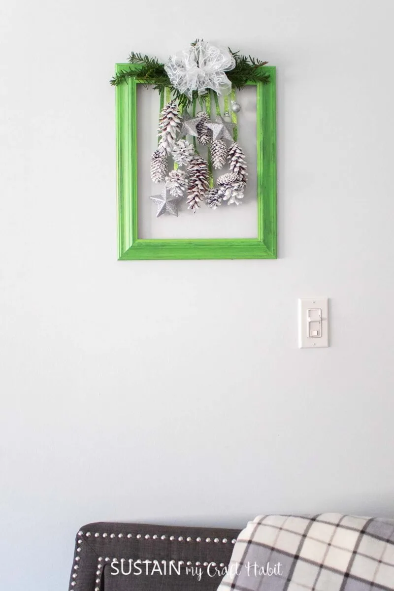 Upcycled picture frame decor for the holidays with greenery, pine cones and ornaments hanging on the wall.