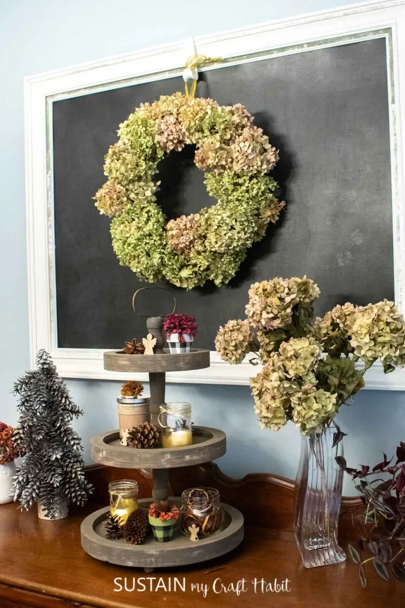 DIY dried hydrangea wreath hung on a chalkboard next to pine cone trees and 3 tiered tray of decor.