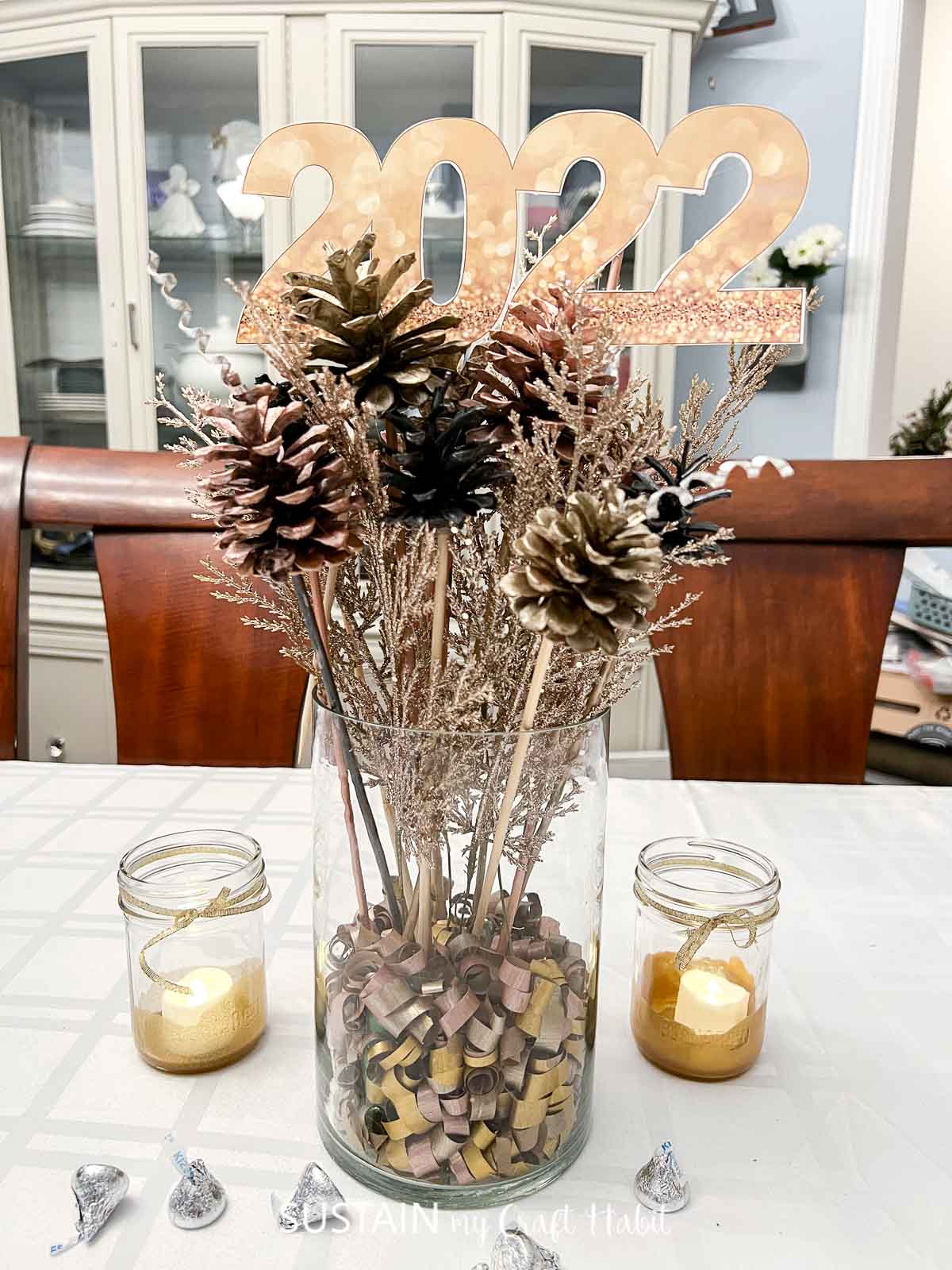 A centerpiece on a dining room table. The centerpiece is made with pine cones painted in gold, rose gold and black metallic colors. A gold 2020 sign is on the top.
