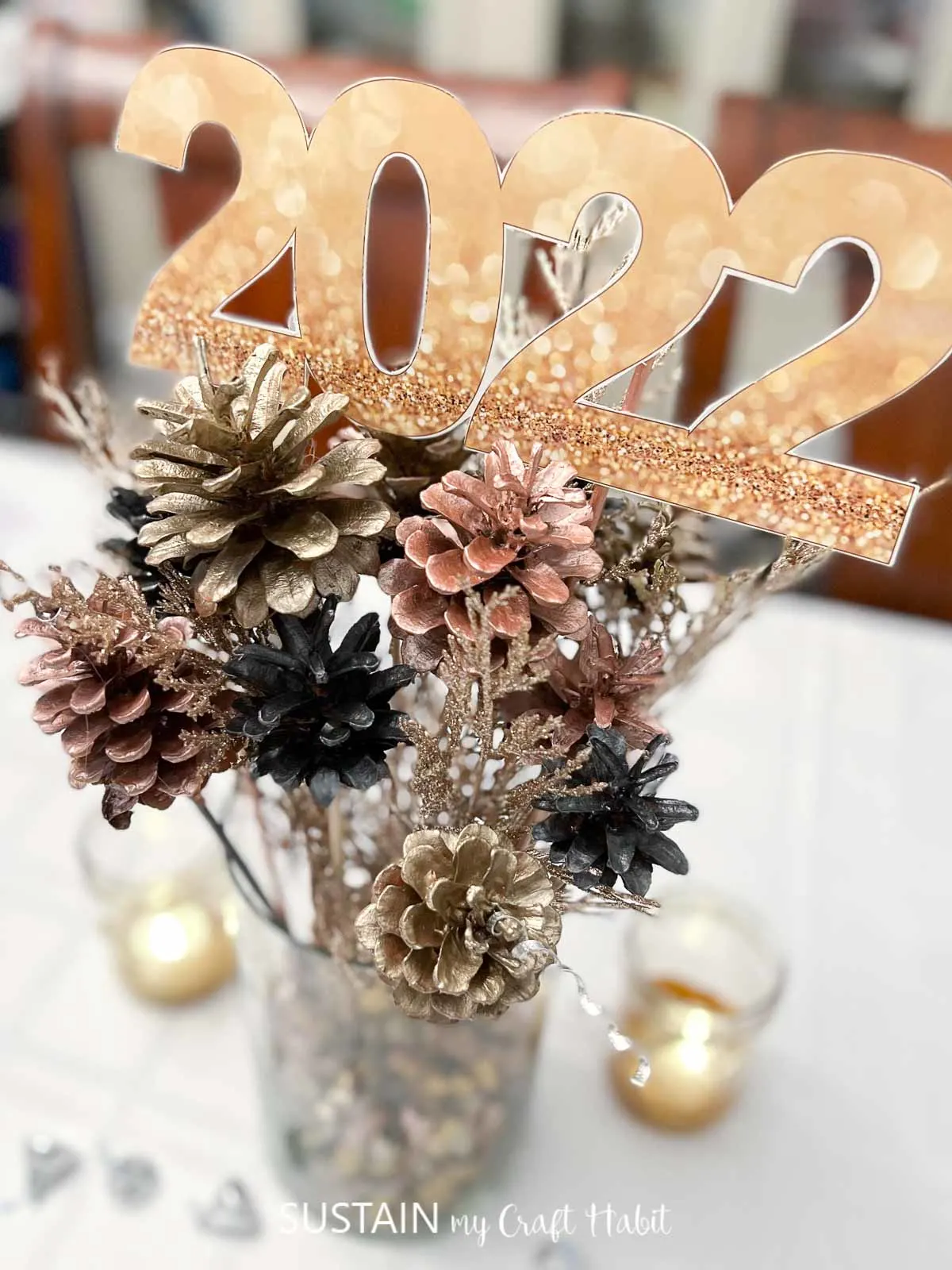 Close up image of the rose gold, gold and balck metallic painted pine cones in a glass vase. A 2022 cut out from the printable is perched on top of the arrangement.