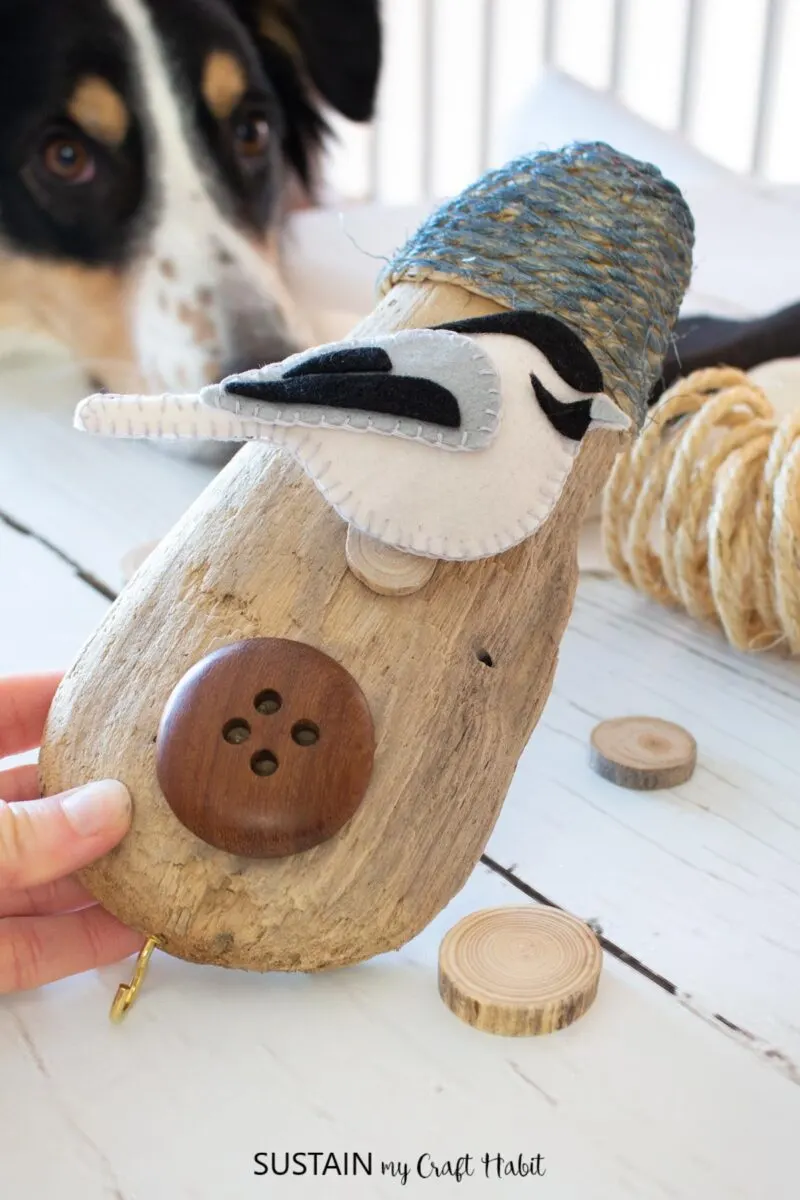 Hand holding birdhouse art craft with driftwood, rope, wood button and a felt bird.