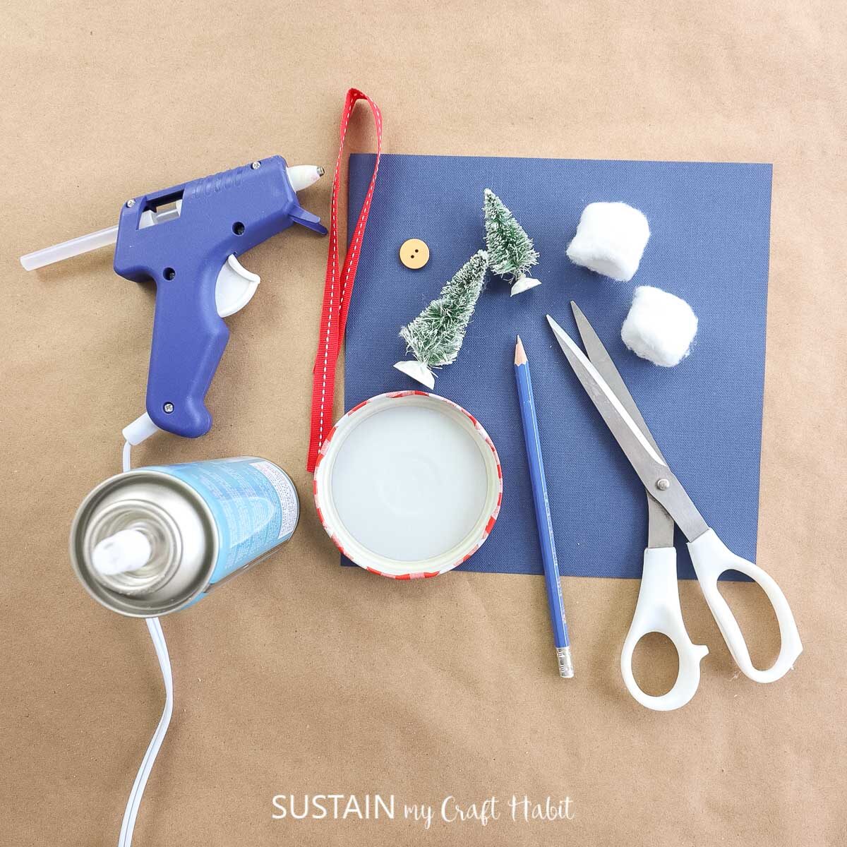 Materials needed to make a bottle brush jar lid ornament including a jar lid, trees, faux snow, ribbon, cotton balls, scissors, pencil and hot glue.