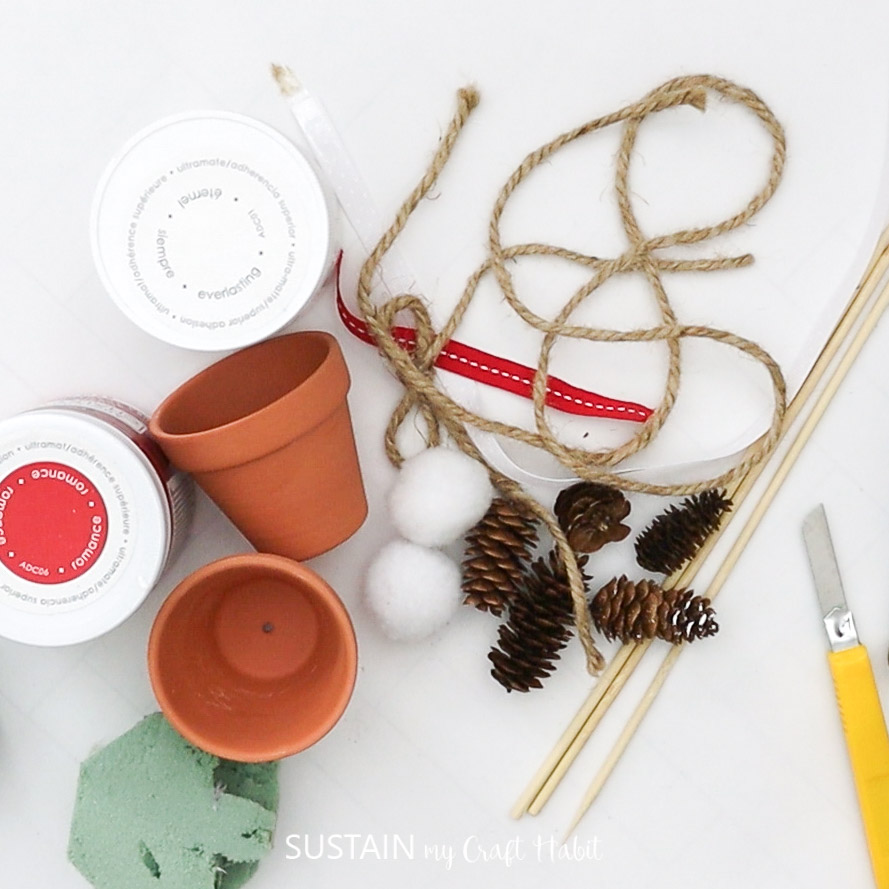 Materials needed to make a mini pinecone topiary craft including pinecones, clay pots, paint, twine, wooden dowels, ribbon, cotton balls, foam and knife.