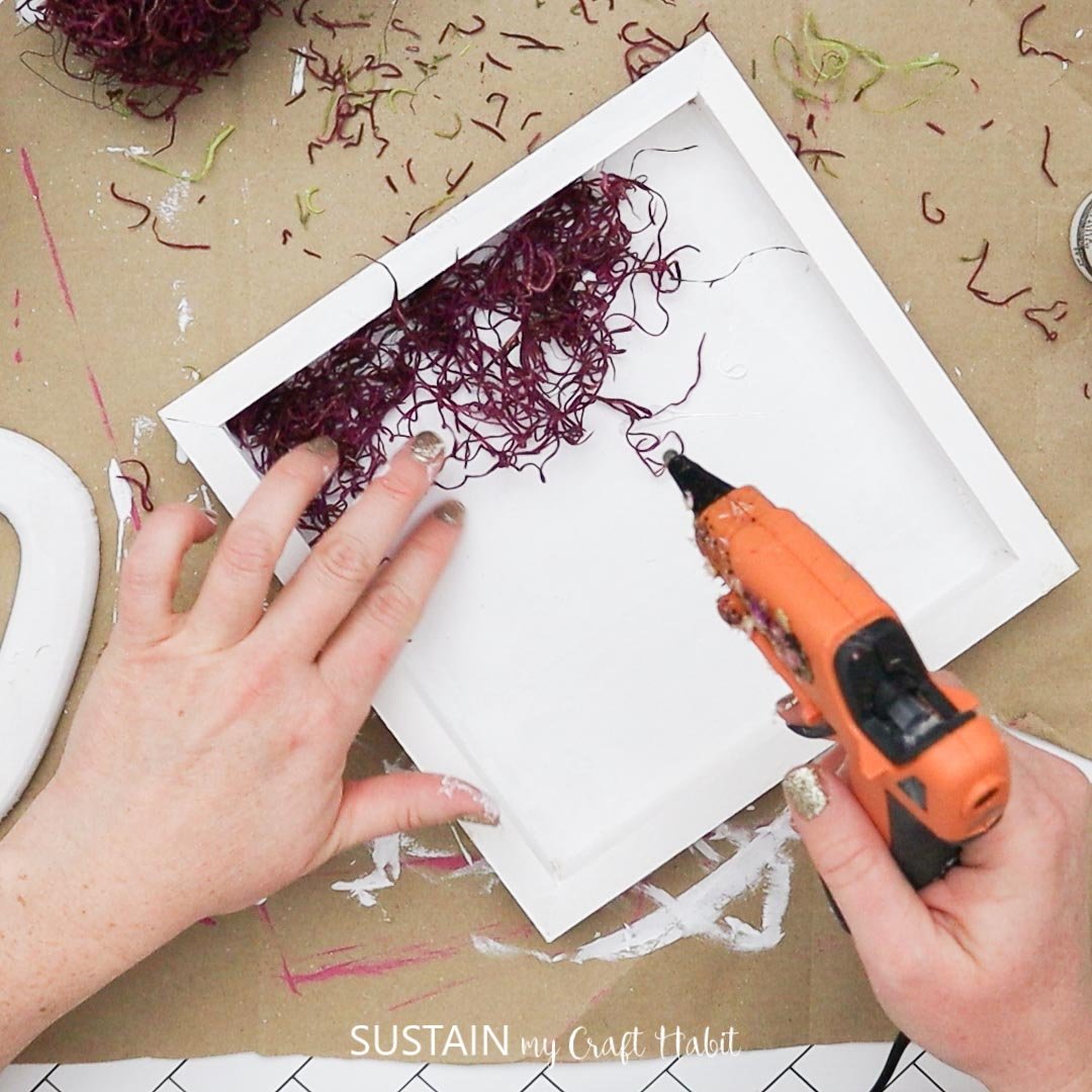 Hot gluing purple moss into the painted wood plaque.