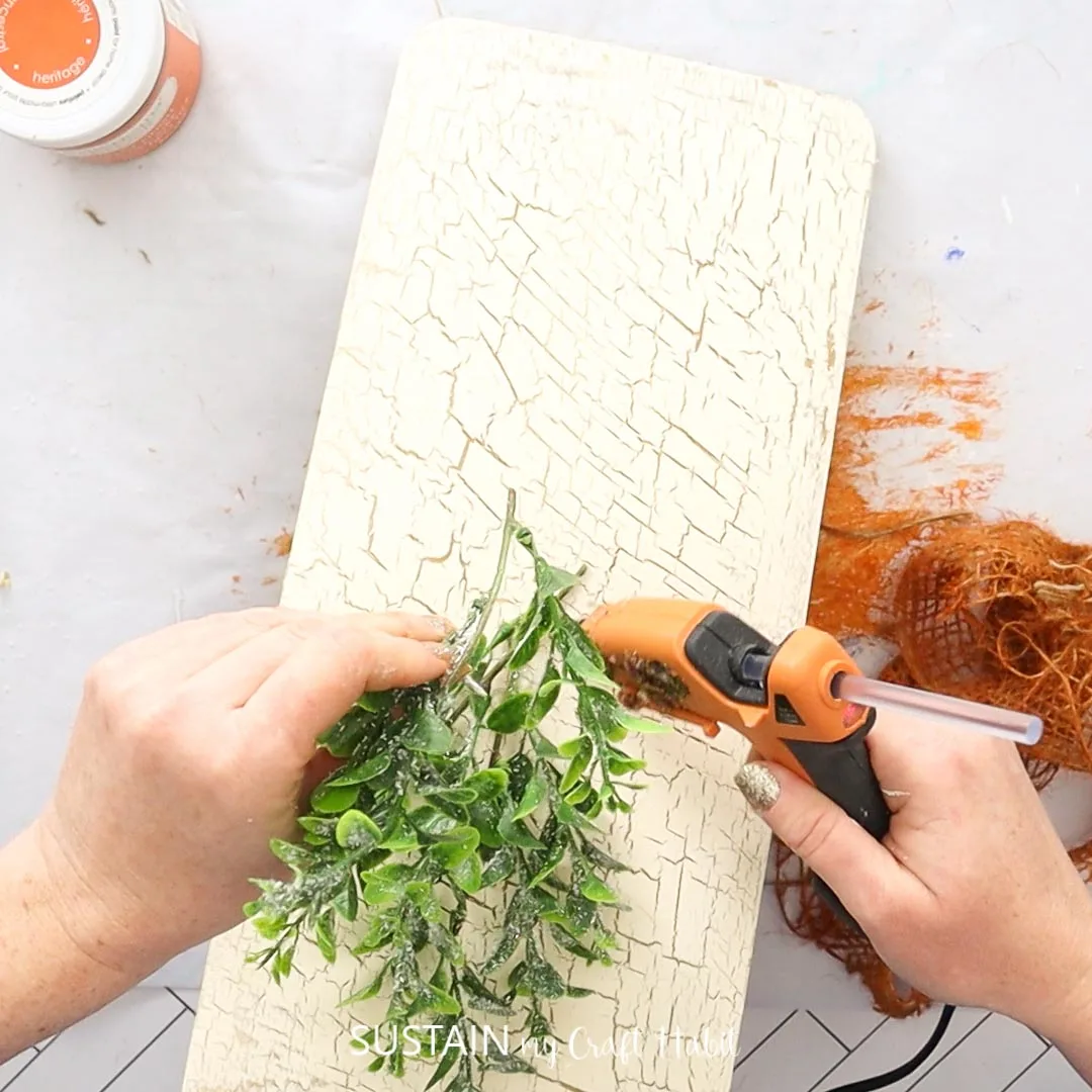 Hot gluing faux greenery onto a painted cedar plank.