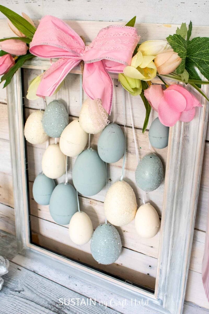 Painted plastic Easter eggs hung with ribbon onto an upcycled picture frame decorated with with faux tulips and a bow.