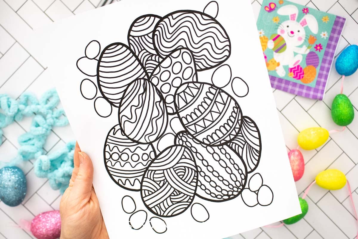 Hand holding an Easter egg coloring page next to Easter eggs and bunny decor.