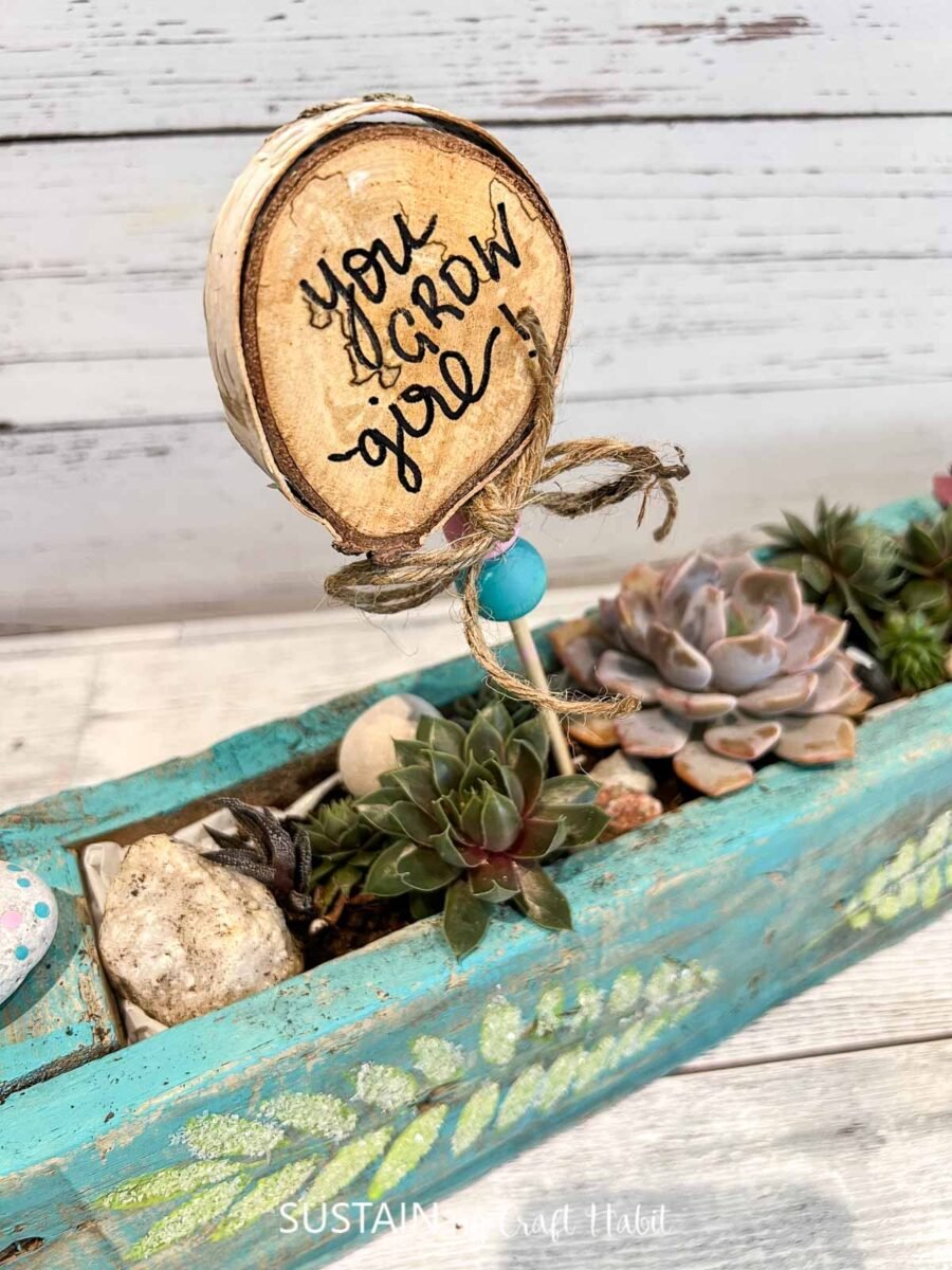 Painted succulent driftwood planter filled with rocks, succulents and a wood slice sign.