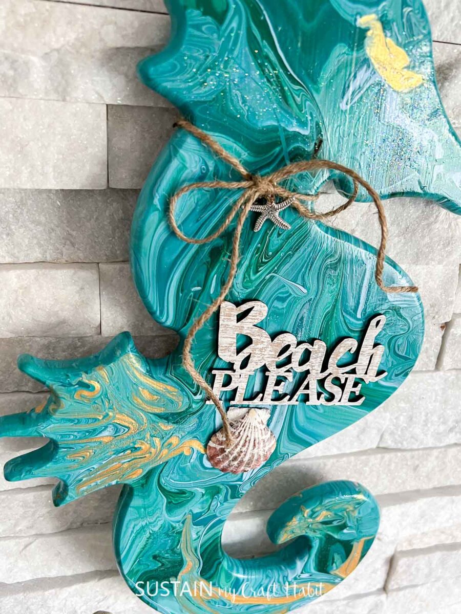 Close up of a paint poured wooden seahorse embellished with a wood cut out phrase, twine, charms and a seashell. 